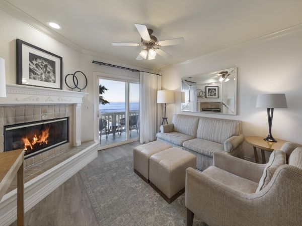 Cozy living room with a built-in fireplace, beige sofa set, ottomans, a large mirror, and a balcony overlooking the water, bright and inviting.