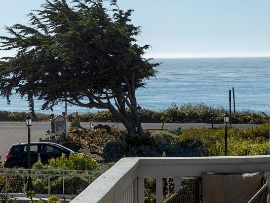 A balcony view overlooking a fence, trees, and the ocean beyond, with a clear sky and a side of a building visible.