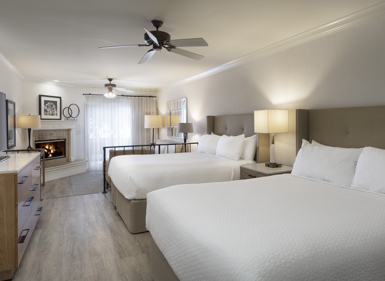 A hotel room with two double beds, a fireplace, lamps, and a ceiling fan.