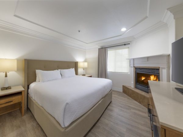 A cozy hotel room with a king bed, two nightstands, soft lighting, a fireplace, and a desk workspace near a window&mdash;calm, modern decor.