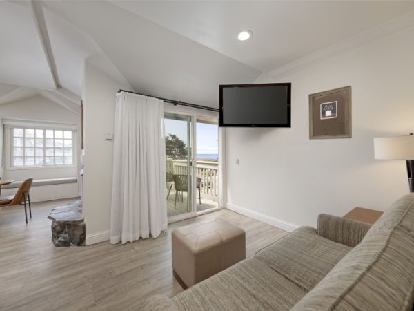 Cozy living room with light gray sofa, wall-mounted TV, large sliding door to balcony, white curtains, and a view into a bright dining area.