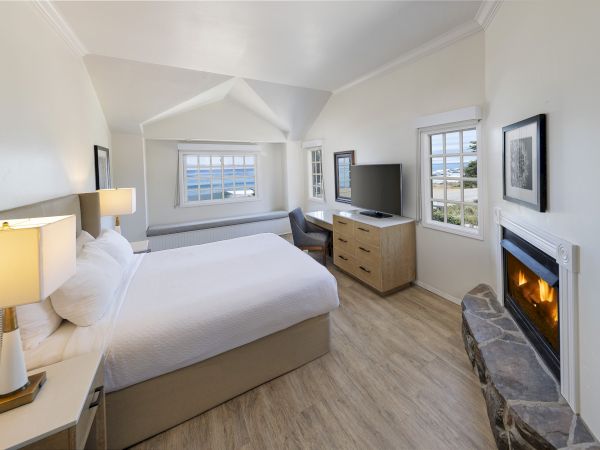A bright hotel room with a king bed, a desk by the window, a dresser with a TV, and a stone fireplace beside a cozy seating area.