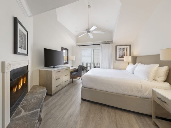 A bright hotel-style bedroom with a king bed, fireplace, desk, flat-screen TV, ceiling fan, and a balcony door at the far end.