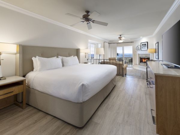 A spacious hotel room with a large bed, nightstands, a desk with a TV, and a seating area by the window, all in light wood tones and neutral decor.