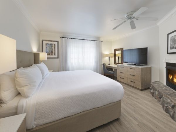 A bright hotel-style bedroom with a large bed, fireplace, flat-screen TV on a dresser, a window with sheer curtains, and a ceiling fan.