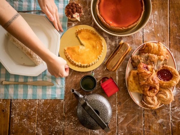 A rustic table with pies, pastries, jam, and a person slicing a pie; cozy baking scene with a kettle and croissants. End sentence.
