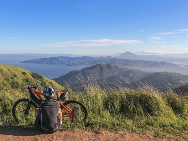 A loaded red touring bike rests by tall grass on a cliff edge, backpack and gear packed, overlooking rolling green hills and a distant blue sea under a clear sky.