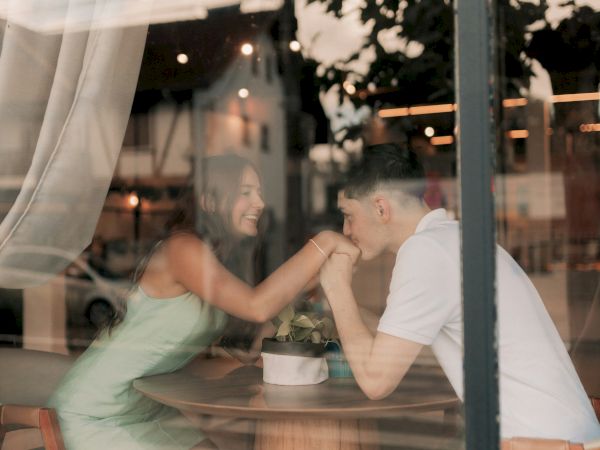 Two people sit at a table in a cafe, smiling and touching foreheads, seen through a window with curtains and reflections.