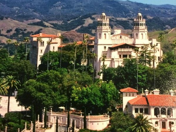 A sunny hillside city with palm trees, historic buildings, and a scenic backdrop of mountains and blue sky.