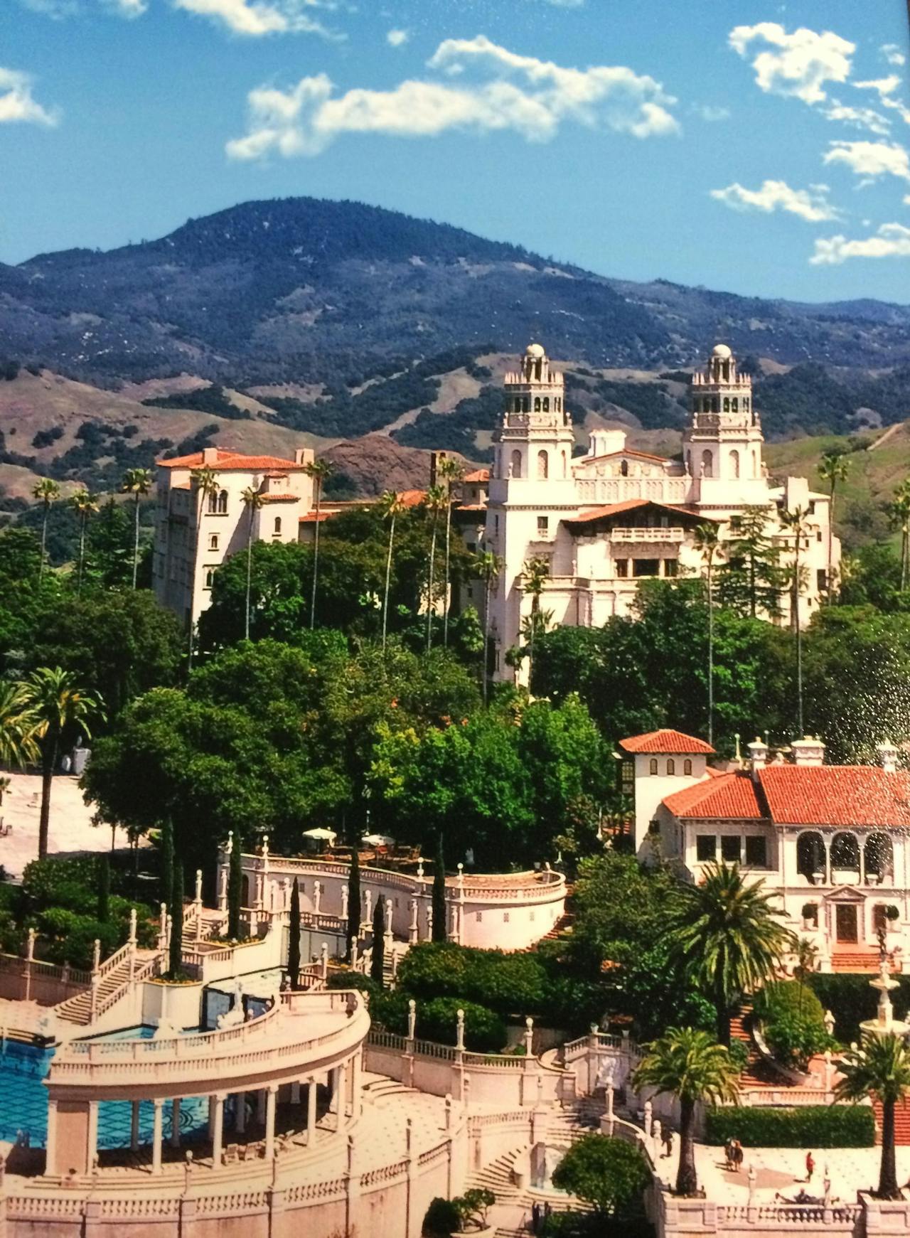 A large white building with red roofs and towers surrounded by trees and a pool