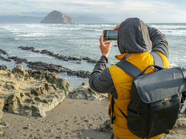 A person with a yellow jacket and backpack is photographing a rocky shore and an island on the horizon at the beach.