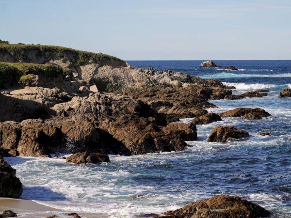 A rocky coastline with waves crashing against jagged rocks along the shore, under a clear blue sky.