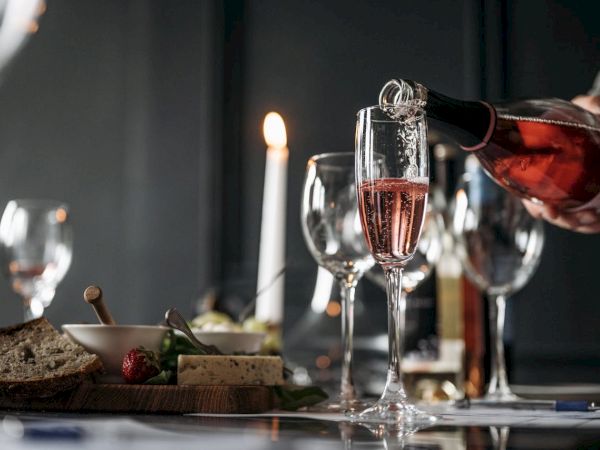 A person pours rose wine into a flute as a candlelit meal with cheese, bread, and glasses is set on a dark dining table.
