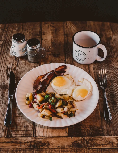 A rustic breakfast: a plate with sunny sides, potatoes and peppers, two fried eggs, crispy bacon, on a wooden table with coffee mug.