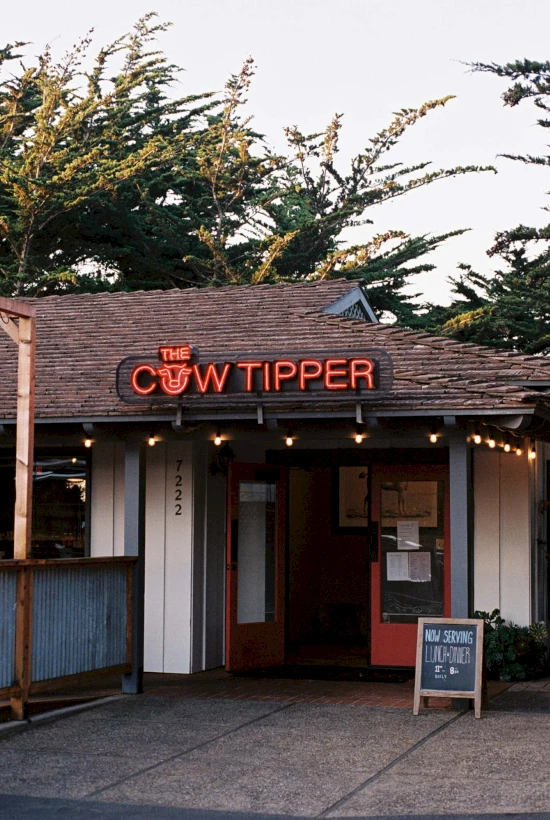 A small cafe or bar named "The Cow Tipper" with a wooden storefront, neon sign, and a chalkboard outside. Ending the sentence.