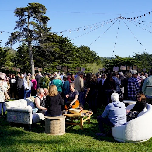A crowded outdoor gathering with people mingling, string lights overhead, and casual seating on the grass, likely a social event or party.