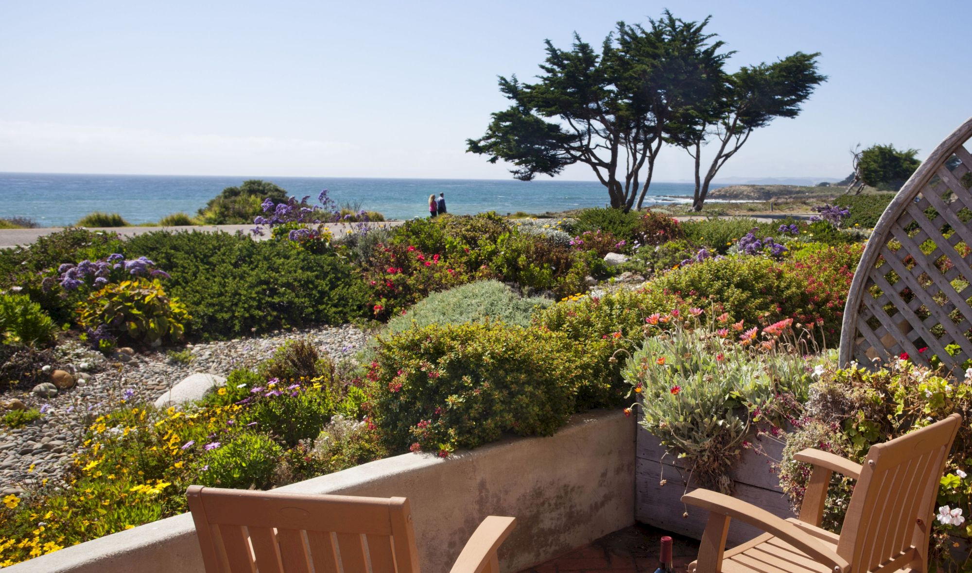 A sunny coastal terrace with two wooden chairs and a small table overlooking lush shrubs and the sea, framed by a lattice screen.