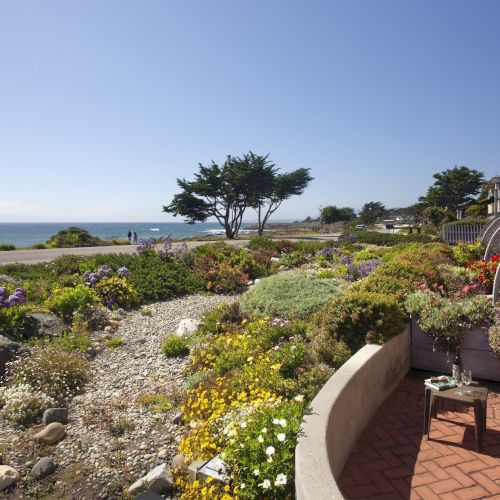 A coastal garden view with colorful flowers, a rocky path, a seawall, and a patio with chairs facing the ocean under a clear sky.