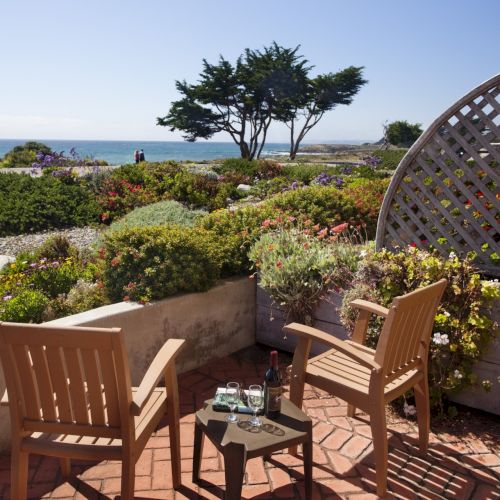 A sunny balcony with wooden chairs and a small table, overlooking a coastal garden and the sea beyond, with a lattice screen on the right.