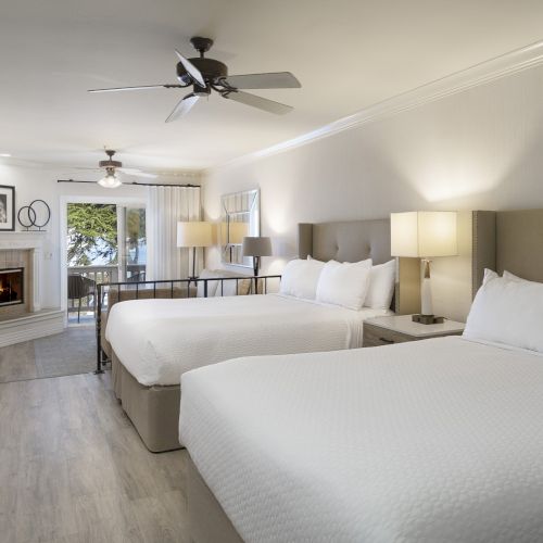 A modern hotel room with two large beds, a desk, flat-screen TV, a seating area near a fireplace, warm lighting, and a balcony view.