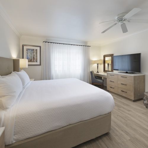 A bright hotel-style bedroom with a large bed, nightstands, a desk, a flat-screen TV, a fireplace, neutral decor, and a stone fireplace surround.