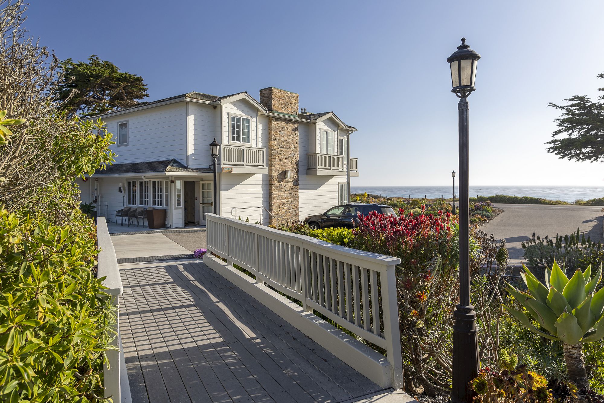 A seaside two-story house with a wooden ramp, white fencing, a lamp post, and a sunny coastal view in the background.