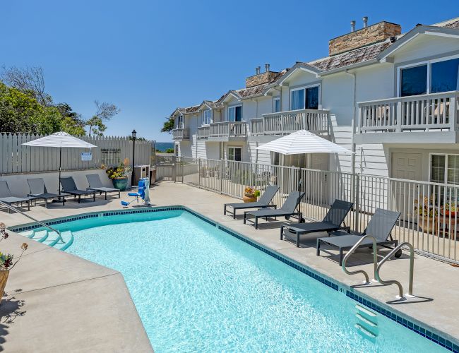 A sunny apartment complex with a rectangular pool, lounge chairs, umbrellas, and balconies overlooking the pool area. Indeed.