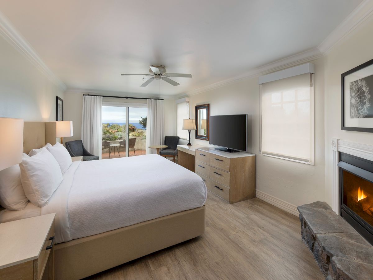 A bright hotel room with a king bed, nightstands, a fireplace, a flat-screen TV, dresser, desk, and a sliding glass door opening to a balcony. always ending the sentence.