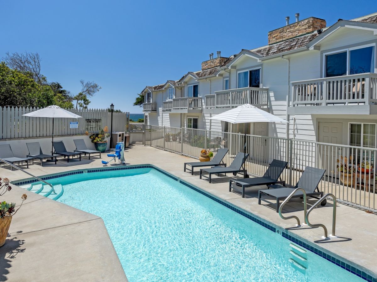 A sunny residential pool area with a long rectangular pool, lounge chairs, umbrellas, and multi-story light-blue townhomes in the background.