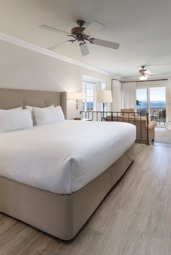 A spacious hotel room with a large king bed, nightstands, seating area, desk, flat-screen TV, wood floors, ceiling fans, and soft lighting.