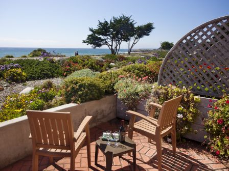 A sunny terrace with wooden chairs and a small table overlooking a coastal garden, ocean in the background, lattice wall on the right.