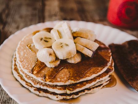 Stack of fluffy pancakes topped with sliced bananas, syrup drizzle, and a side of toast on a rustic plate. Enjoy breakfast vibes.