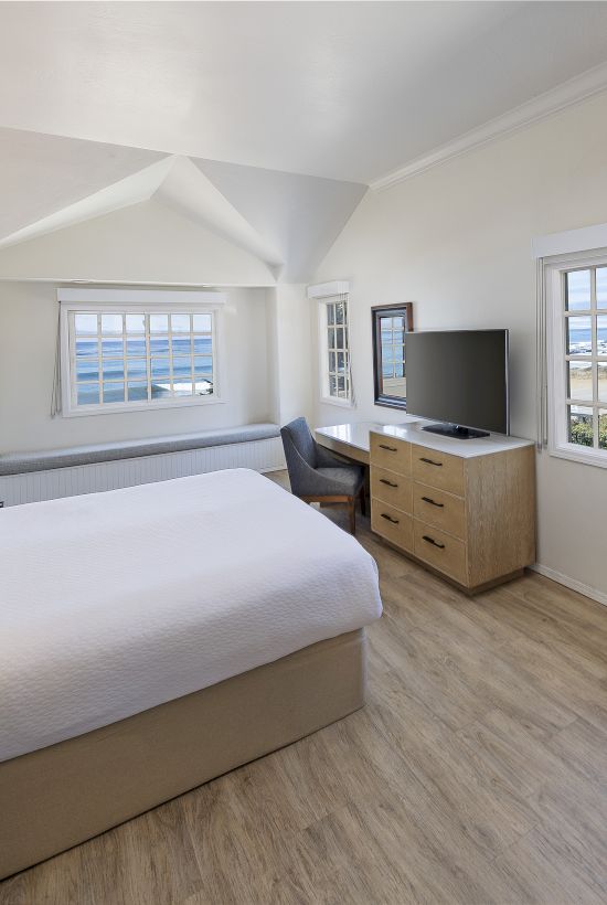 A bright hotel-style bedroom with a large bed, nightstands, a dresser with a TV, a fireplace, and a small seating area by windows in a airy suite.