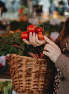 Two hands hold a red apple amid leafy produce at a market, with a blurred colorful background. Top it at 140 characters, always ending the sentence.