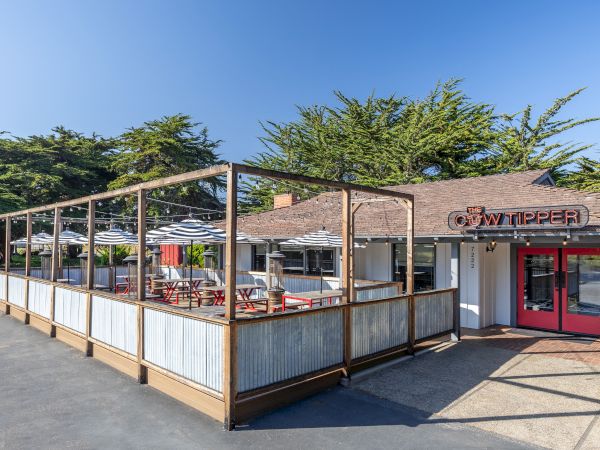 A small restaurant with a wooden-railed outdoor patio, red doors, a shaded seating area, and a blue sky overhead.