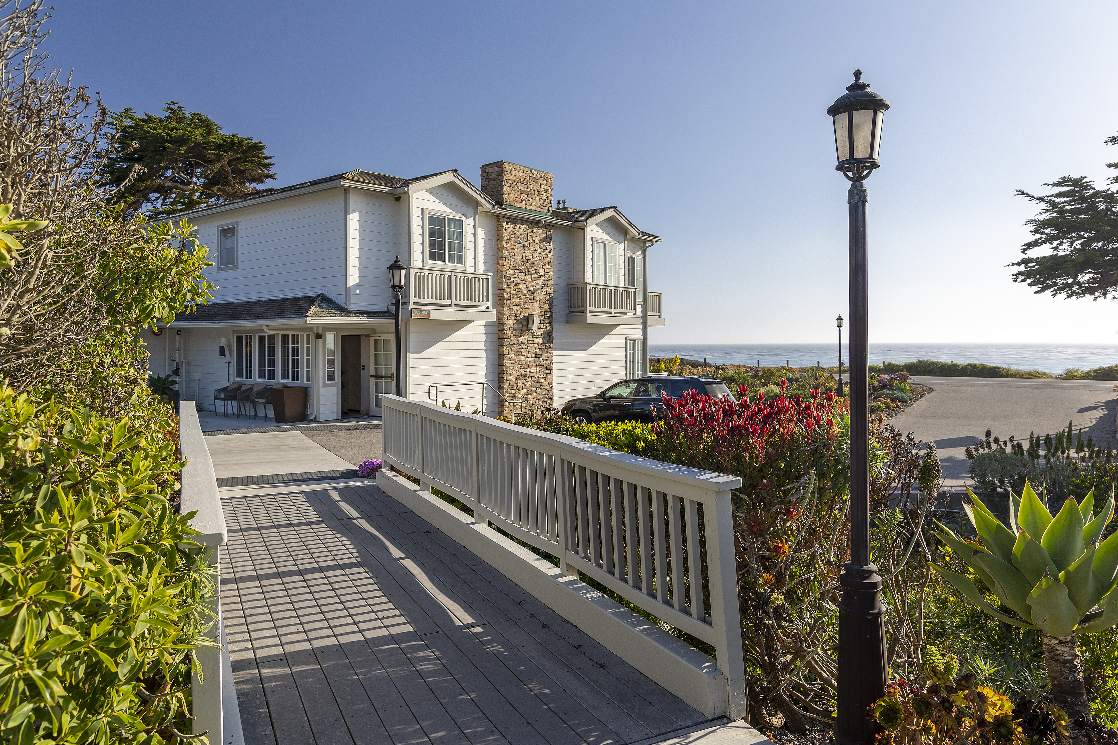 A white building with a balcony and stone chimney, surrounded by plants and a pathway